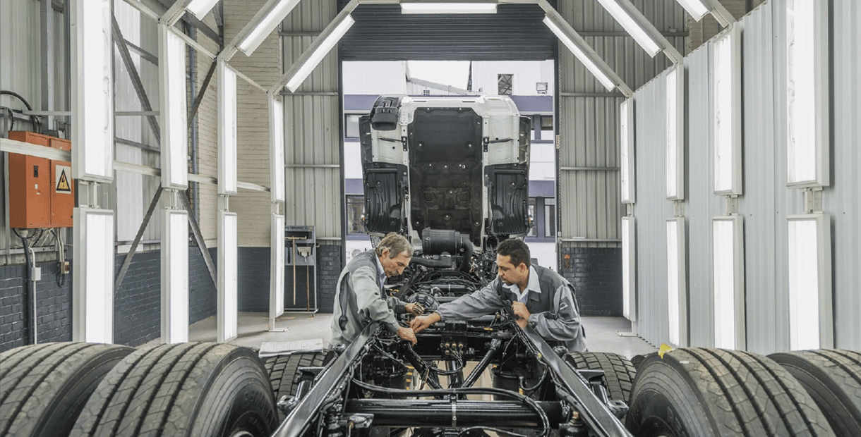 Twee monteurs die in een goed verlichte werkplaats aan het chassis van een vrachtwagen werken, met de cabine omhoog geklapt en grote banden zichtbaar op de voorgrond.