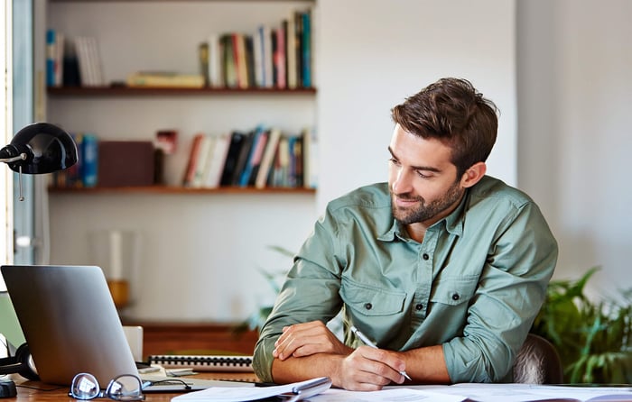 Man thuis kijk naar laptop terwijl hij iets opschrijft.