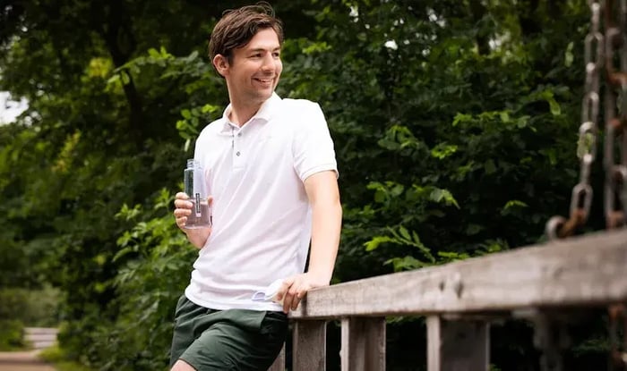 Man in een witte polo en groene broek leunt tegen een houten hek in een groene buitenomgeving, terwijl hij een flesje water vasthoudt en glimlachend opzij kijkt.