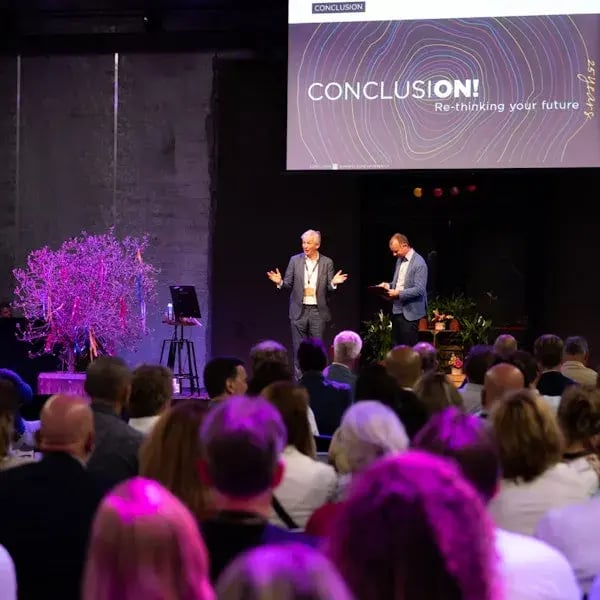 Twee mannen op een podium die een Conclusion presentatie geven aan groot publiek