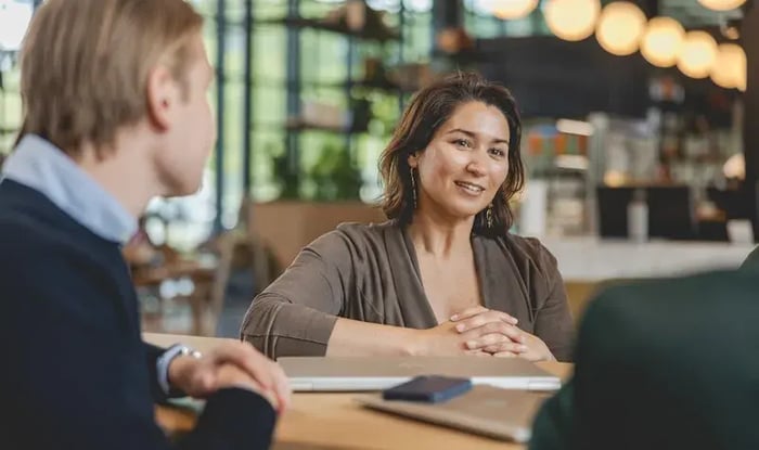 Vrouw zit aan tafel en spreekt tijdens een gesprek met twee anderen in een moderne, groene kantoor- of horecaruimte, met een laptop en smartphone op tafel.