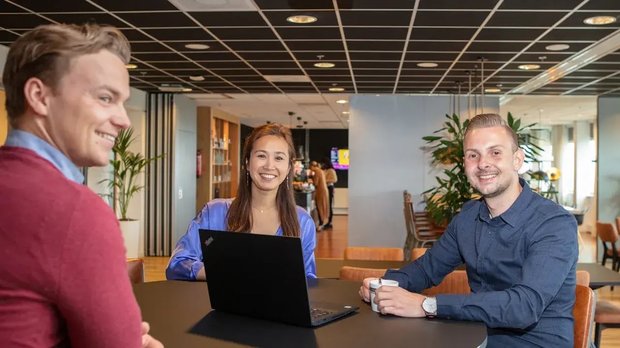Three young employees in the office gathered around their laptop, smiling at the camera.