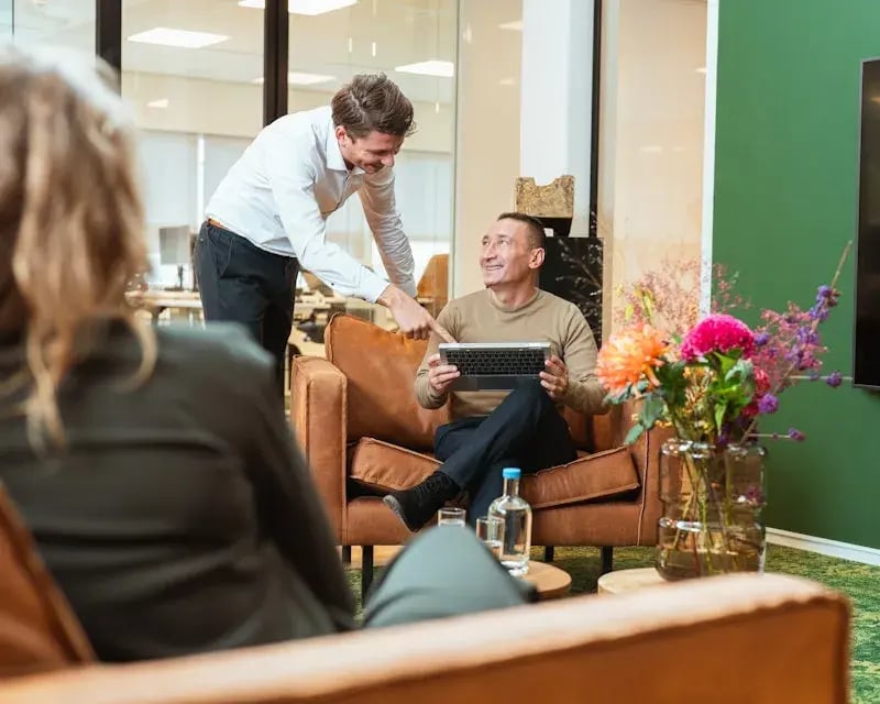 Informele kantoorsetting met een man op een bruine leren bank die een laptop vastheeft en een man in wit overhemd wijst hem wat aan