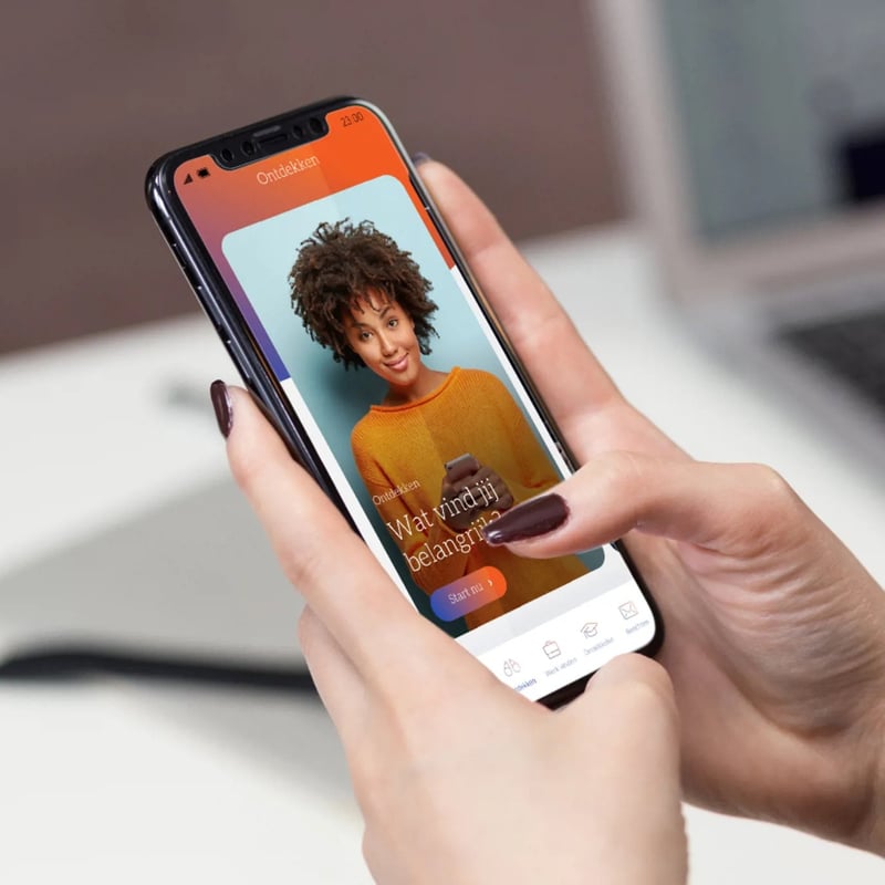 Handen die een smartphone vasthouden met de Olympia-app geopend, waarop een vrouw in een oranje trui wordt getoond.