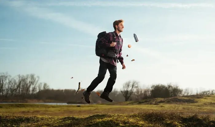 Man met een rugzak die buiten door een open landschap springt of loopt, terwijl bladeren en een waterfles door de lucht bewegen onder een heldere blauwe lucht.