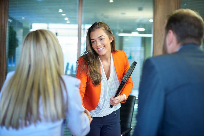 A female colleague in an orange blazer is shaking hands with another woman.