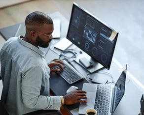A male colleague is behind his desk working on two computers.