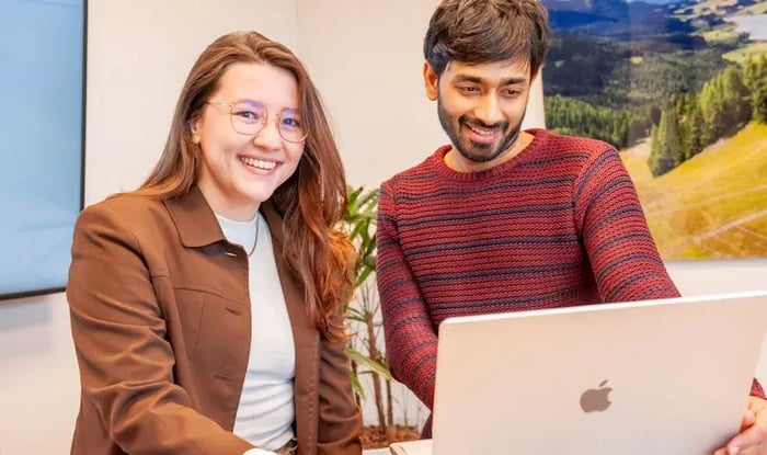 A female colleague is smiling in front of her and a male colleague is smiling at a laptop.