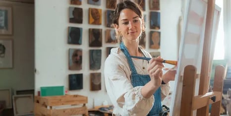 Vrouw in een atelier die geconcentreerd schildert op een doek op een ezel, met portretten aan de muur op de achtergrond.