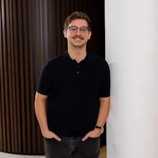 A man, Valentin Calomme, with dark hair, a mustache and glasses in a black polo with hit hands in his pockets, smiling in front of the camera