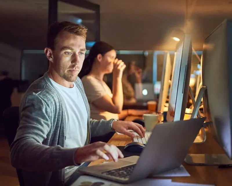 Man op de voorgrond en een vrouw op de achtergrond die allebei in een schemerig kantoorpand op hun laptop en computer werken