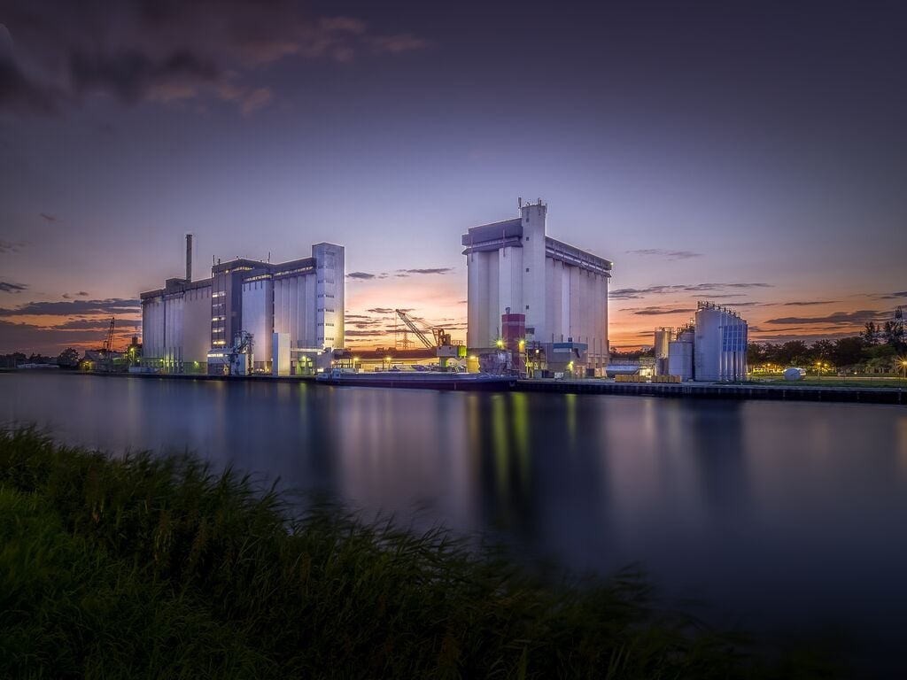 Industrieterrein met grote silo’s en fabriekshallen langs het water bij zonsondergang.