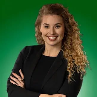 Portret van een glimlachende vrouw met lang krullend blond haar, gekleed in een zwart jasje, met haar armen over elkaar tegen een groene achtergrond.