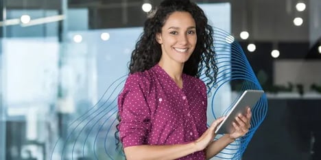 Uma senhora branca de cabelo castanho comprido e encaracolado, sorrindo para a câmara, com uma blusa magenta com bolinhas brancas e um tablet na mão