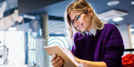 Uma senhora branca, de cabelo loiro, de óculos, com um casaco de malha roxo, a sorrir para o seu tablet