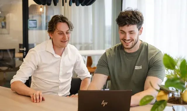 Twee collega's kijken lachend naar een laptop.