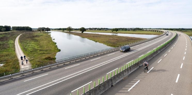 Snelweg en fietspad door een waterrijk Nederlands landschap, met een auto en meerdere fietsers onderweg tussen groene oevers en open velden.