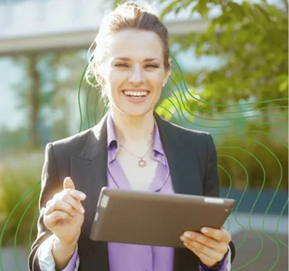 Uma mulher branca com cabelo escuro preso em um rabo de cavalo, vestindo uma camisa roxa, sorrindo para a câmera com um tablet na mão