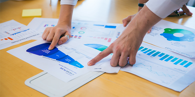 Twee personen wijzen naar grafieken en diagrammen op papieren rapporten tijdens een data-analyse.