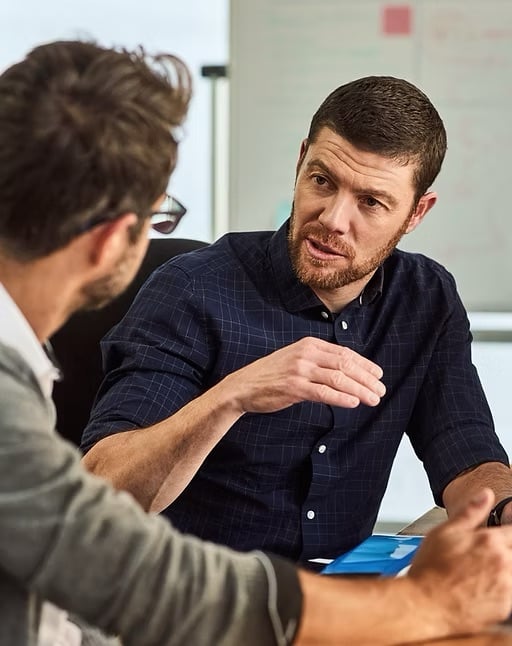 Twee mannen in gesprek aan een vergadertafel, met een whiteboard op de achtergrond.