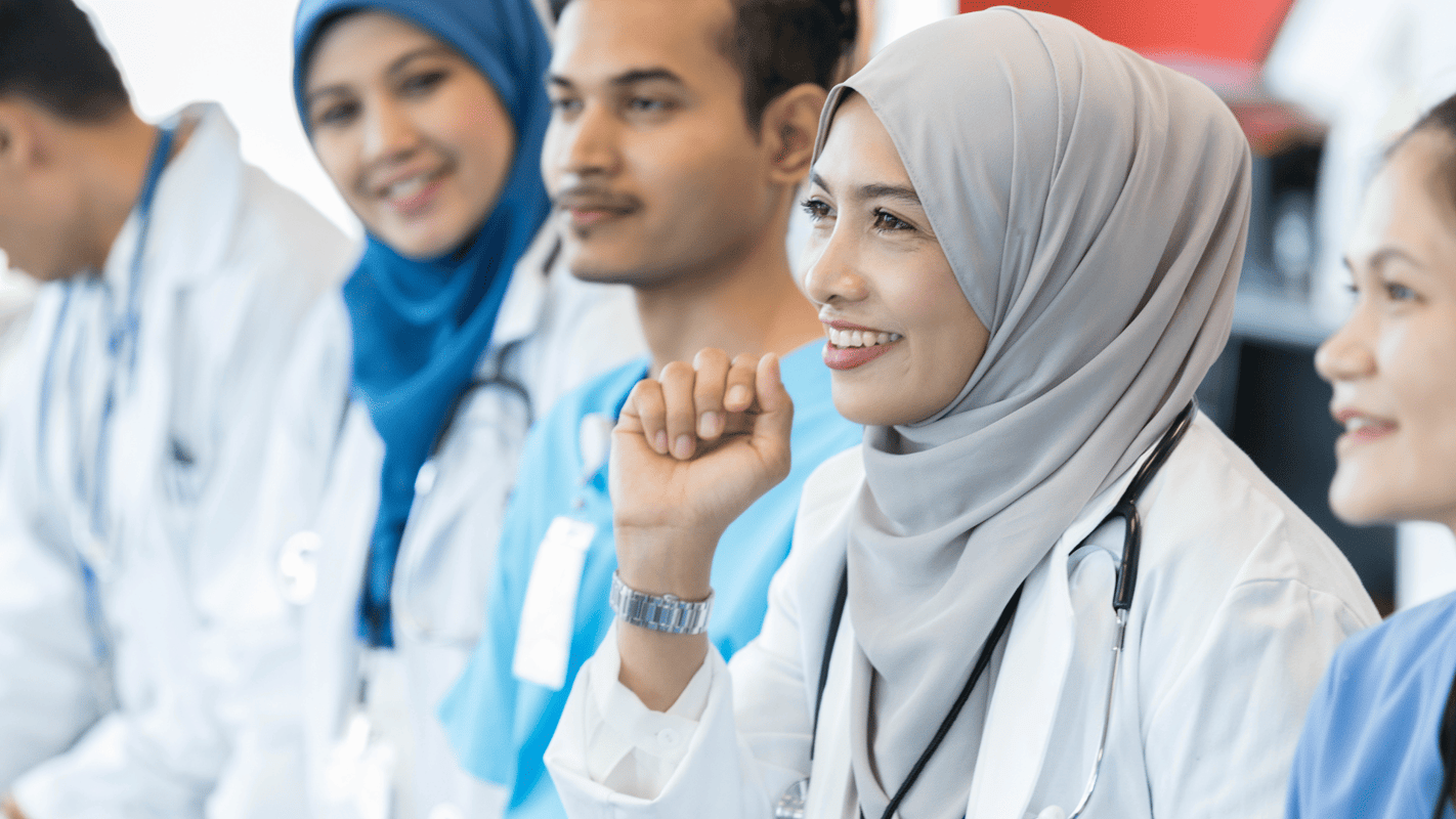 Verpleegers zitten naast elkaar, eentje met een hoofdoek heeft een vraag