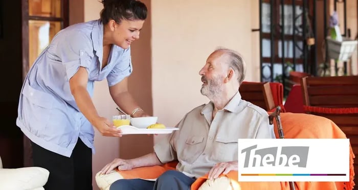 Een verpleegster geeft een man zijn ontbijt met de Thebe logo in een hoekje