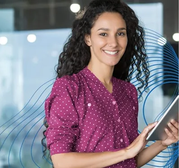 Uma mulher branca com cabelo castanho, longo e encaracolado, sorrindo para a câmera, usando uma blusa magenta com bolinhas brancas e segurando um tablet na mão.