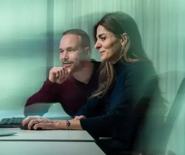Two colleagues behind a desk are smiling at their computer.