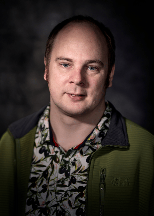 Een portret van een man met een groene jas en een bloemenprint overhemd. Hij heeft kort bruin haar, blauwe ogen en een lichte glimlach, met een neutrale achtergrond