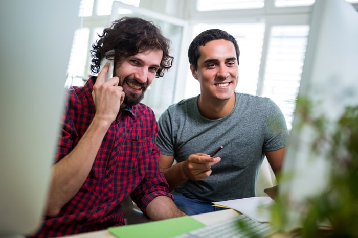 male-graphic-designer-talking-mobile-phone-while-coworker-looking-computer