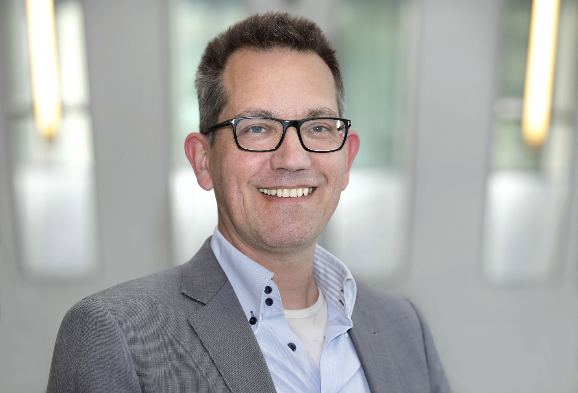 Een man met kort haar, bril en een grijs colbert over een lichtblauw overhemd. Hij glimlacht vriendelijk en staat in een lichte, moderne binnenruimte met een onscherpe achtergrond.