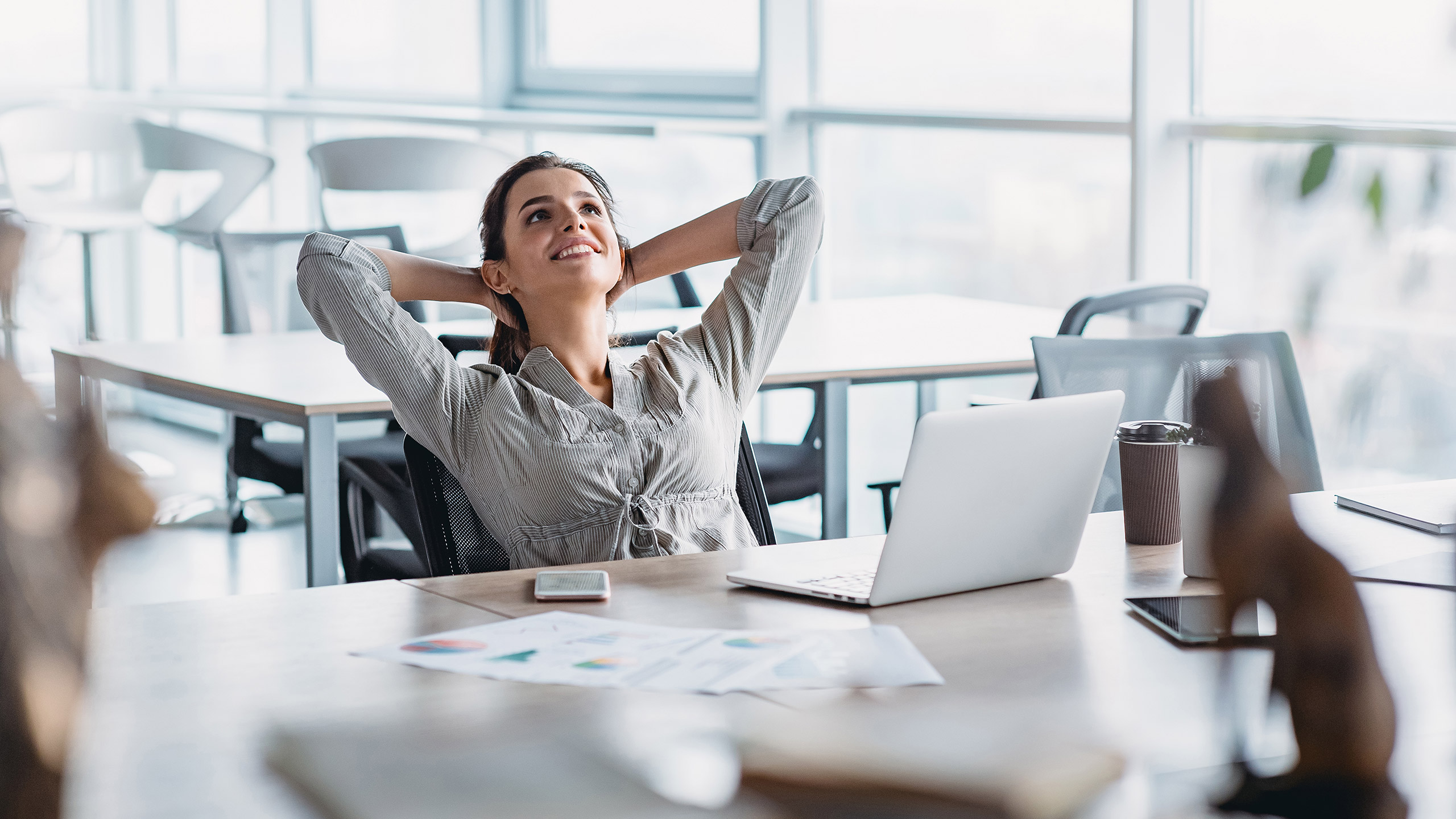 Vrouw zit ontspannen achter haar bureau met laptop, kijkt glimlachend omhoog met handen achter haar hoofd.