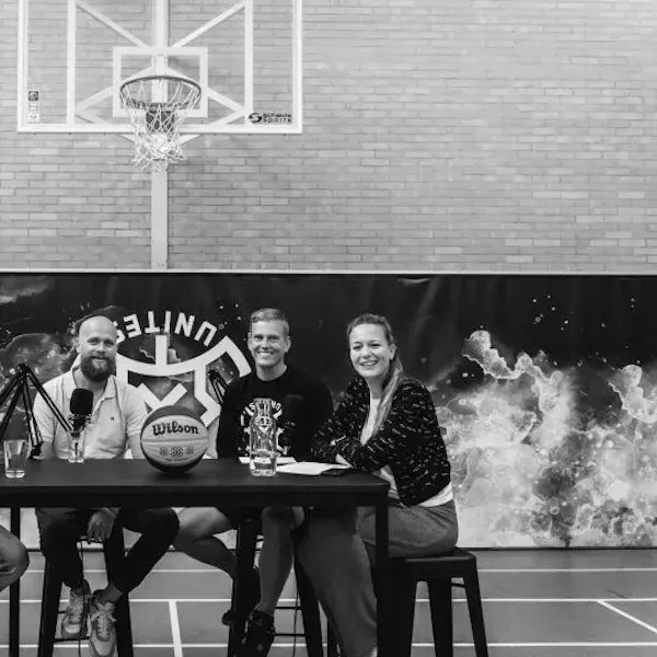 Drie mensen zitten aan een tafel in een sporthal, met microfoons en een basketbal op tafel, terwijl ze poseren voor een opname onder een basketbalbord.