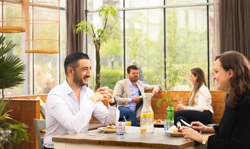 Twee mensen zitten aan een tafel in een lichte, groene horecaomgeving en praten tijdens het eten, terwijl op de achtergrond andere gasten ontspannen zitten tussen planten en grote ramen.