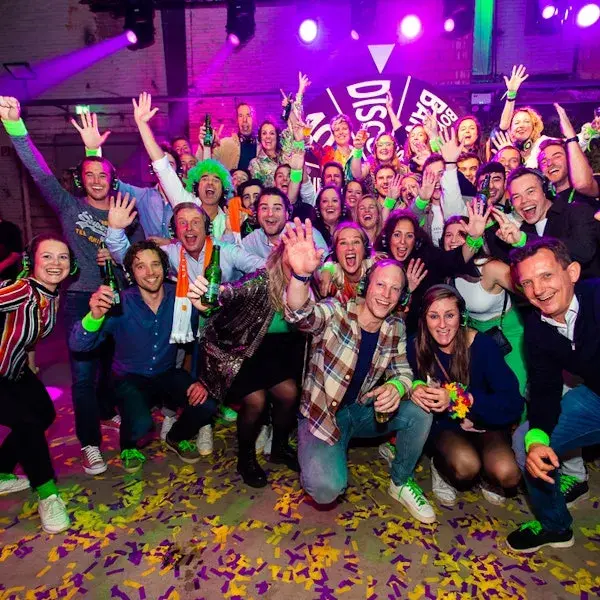 Grote groep mensen viert feest in een kleurrijke, verlichte zaal, met confetti op de vloer; ze lachen, zwaaien naar de camera en houden drankjes vast tijdens een energieke groepsfoto.
