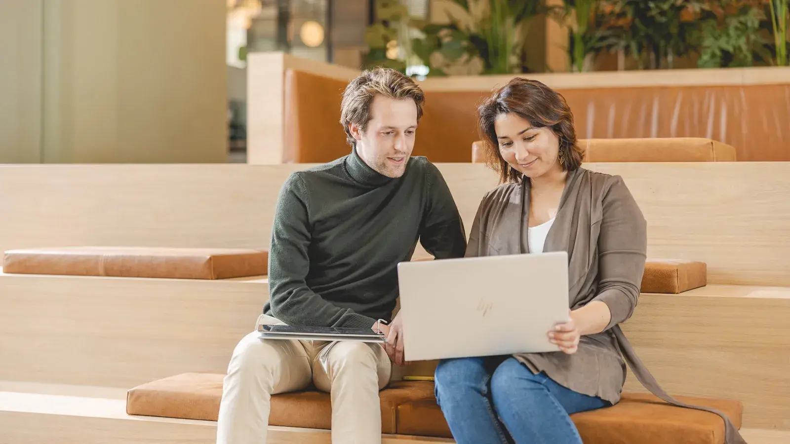 Twee collega’s zitten naast elkaar op houten traptreden in een lichte kantoorruimte en kijken samen naar een open laptop, terwijl ze overleggen.