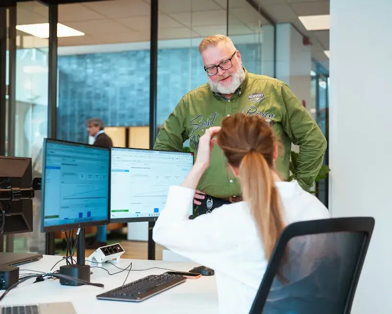Vrouw zit aan bureau met twee beeldschermen en praat met mannelijke collega die naast haar bureau staat