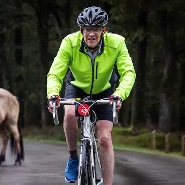 Man in sportieve kleding en fietshelm fietst op een natte weg door een bosrijke omgeving, terwijl het licht regent.