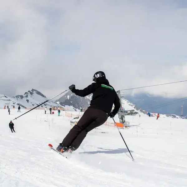 Persoon skiet op een besneeuwde piste in de bergen, gezien van achteren, met skistokken in de handen en andere skiërs en skiliften op de achtergrond.