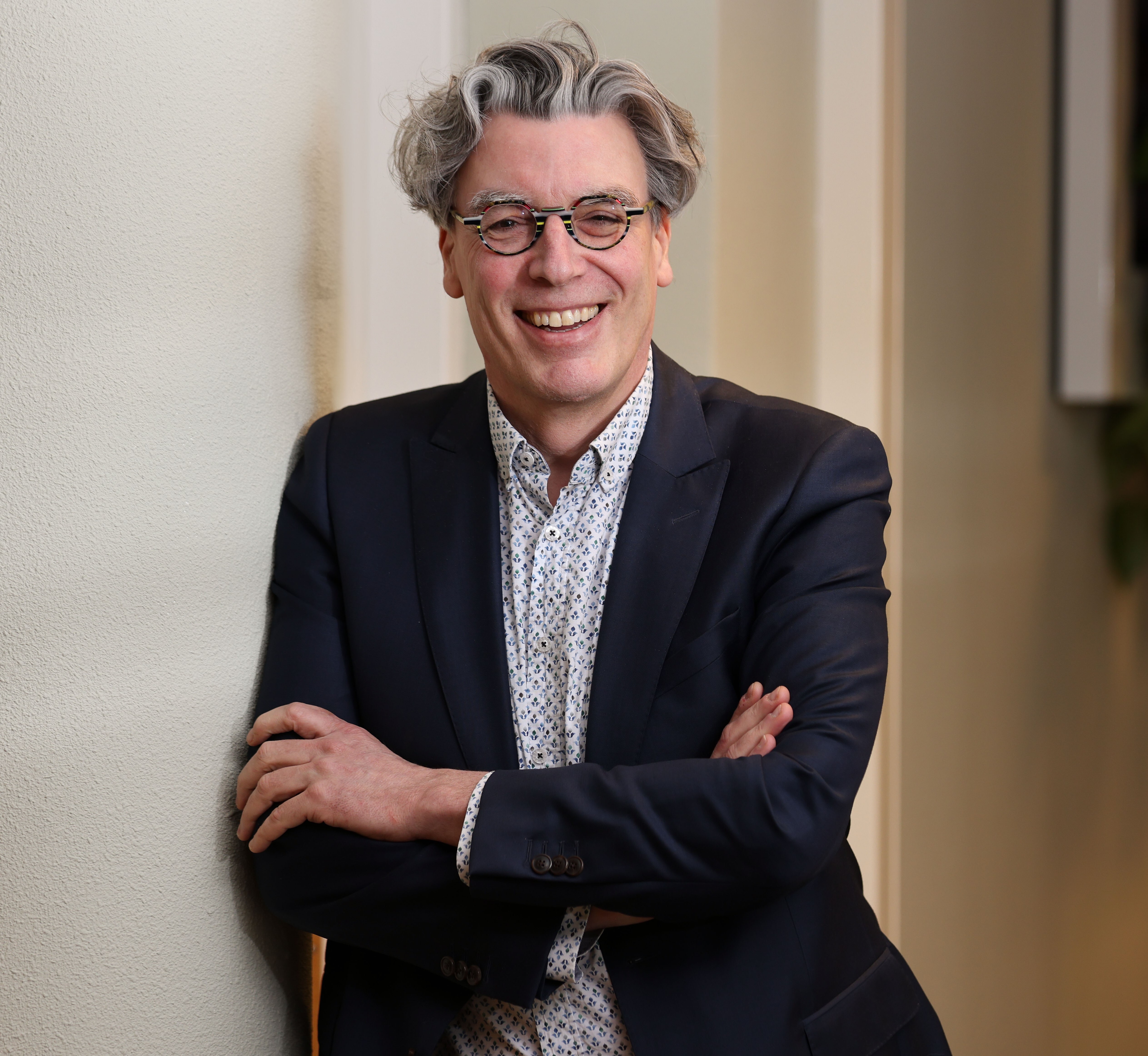  Man with glasses smiles while leaning against a wall with his arms crossed.