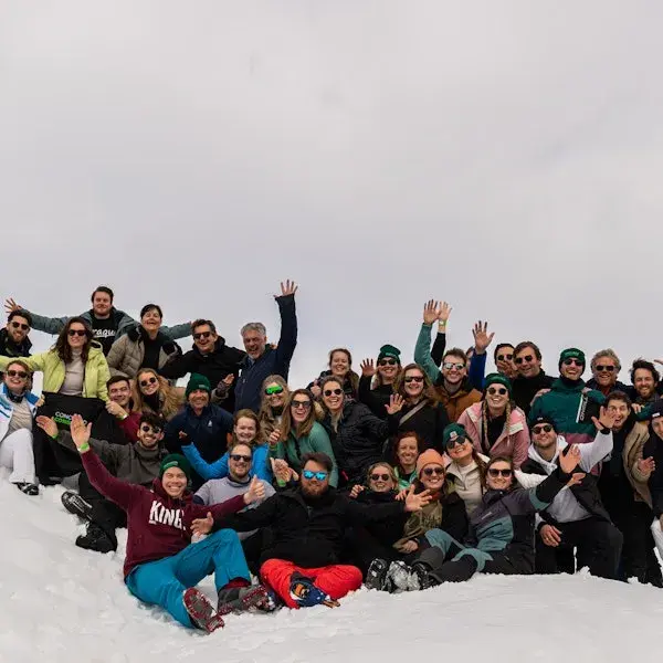 Grote groep mensen in winterkleding poseert lachend in de sneeuw, met opgeheven handen, tijdens een gezamenlijke buitenactiviteit in een bergachtige omgeving.