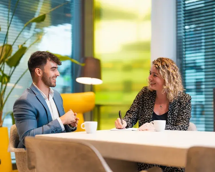 Twee collega's zitten aan een tafel met kopjes koffie en lachen naar elkaar.