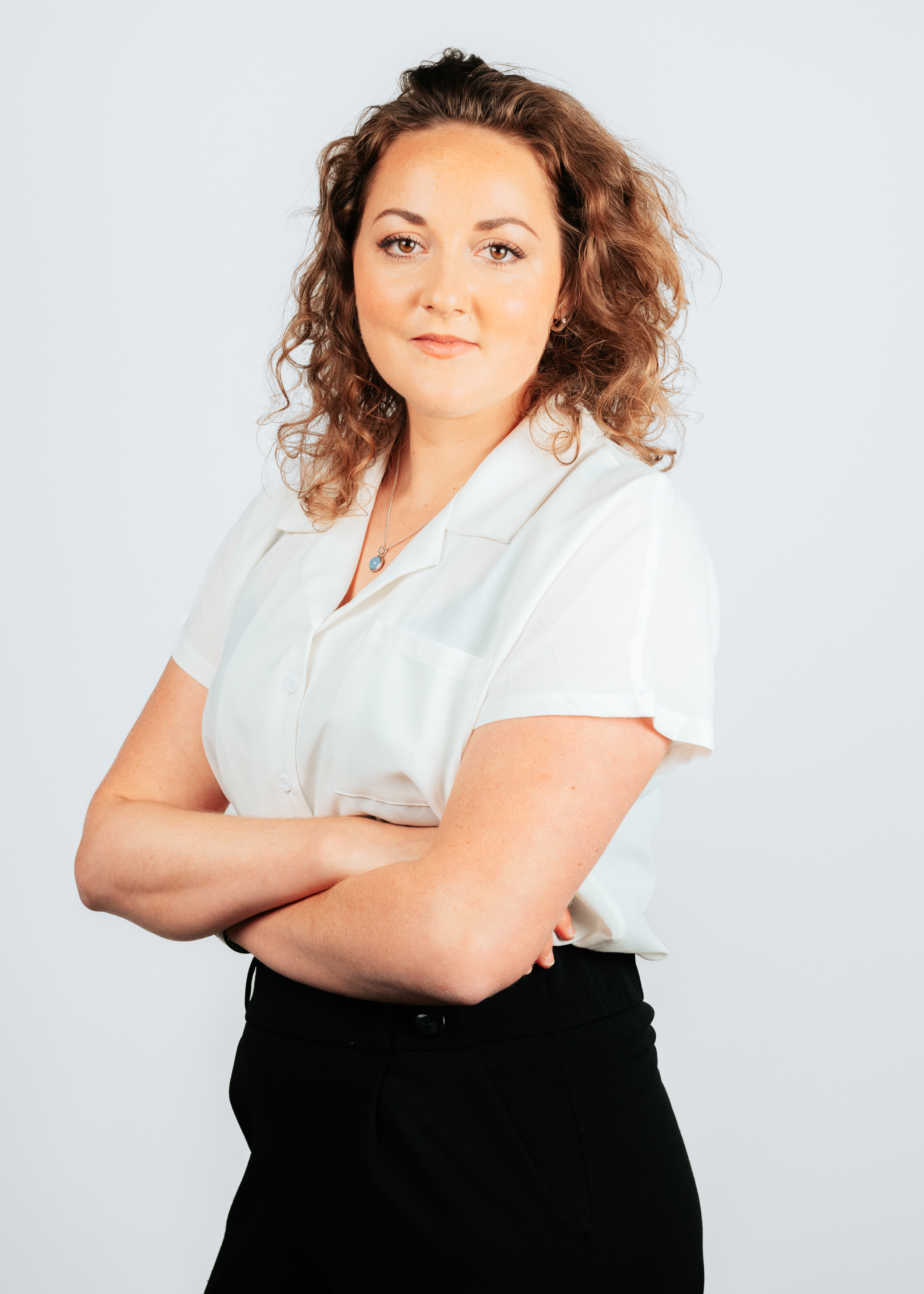 Vrouw met krullend haar die met gekruiste armen poseert, gekleed in een witte blouse en zwarte broek, voor een neutrale lichtgrijze achtergrond.  Select 78 more words to run Humanizer.