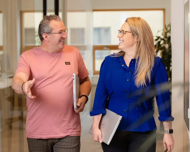 Man en vrouw lopen met hun laptops in de hand lachend door de gangen van het kantoor.