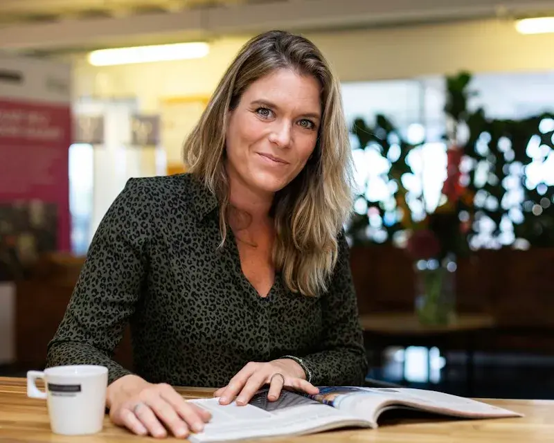 Een dame met een boek en een kop koffie kijkt in de camera